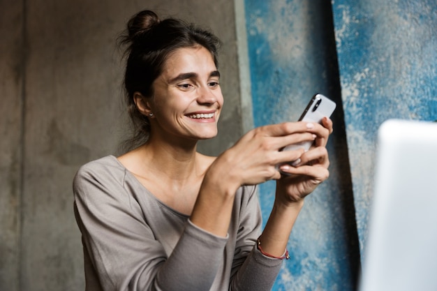 Фото довольно молодой женщины, сидящей в кафе в помещении с помощью портативного компьютера и мобильного телефона.
