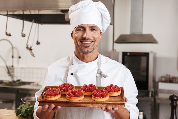 Photo of professional male chief in white uniform holding plate with cakes