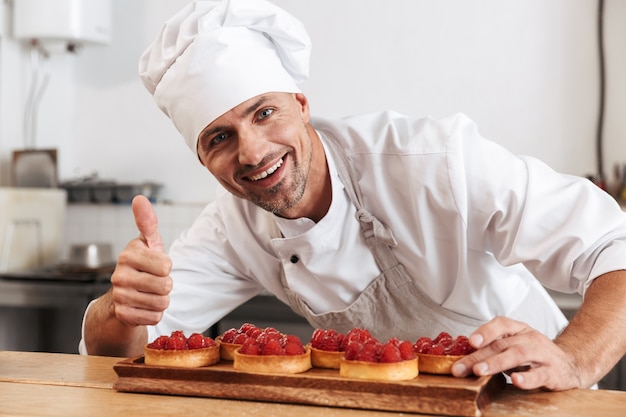 Photo of professional male chief in white uniform holding plate with cakes