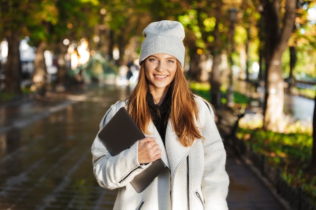 Photo of a redhead happy woman walking outdoors at the street smiling holing laptop computer.