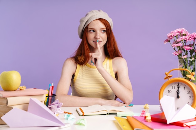 Photo of redhead student girl keeping finger at her mouth while doing homework isolated over purple wall