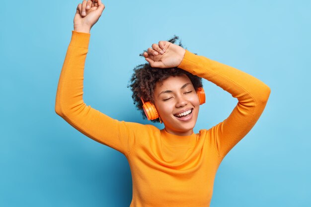 Photo of relaxed curly haired young woman keeps eyes closed dances and catches every bit of music smiles broadly wears casual orange jumper uses wireless headphones isolated over blue wall