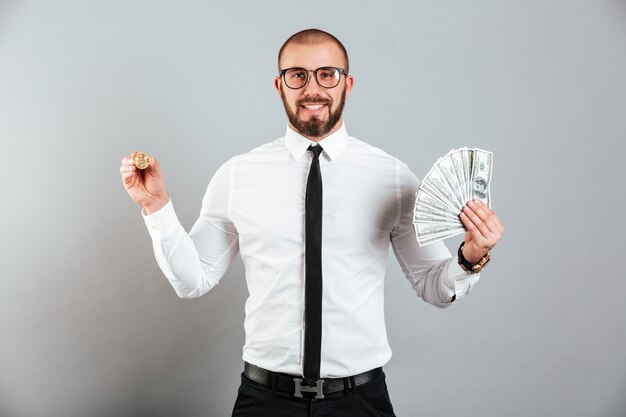 Photo of rich man 30s in glasses and suit showing bitcoin and lots of money dollar currency, isolated over gray wall