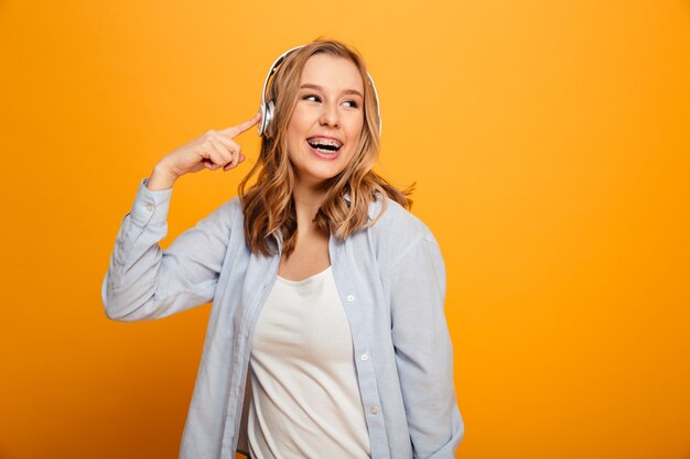 Photo of satisfied smiling woman 20s wearing braces in casual clothing listening to music using wireless earphones, isolated over yellow space