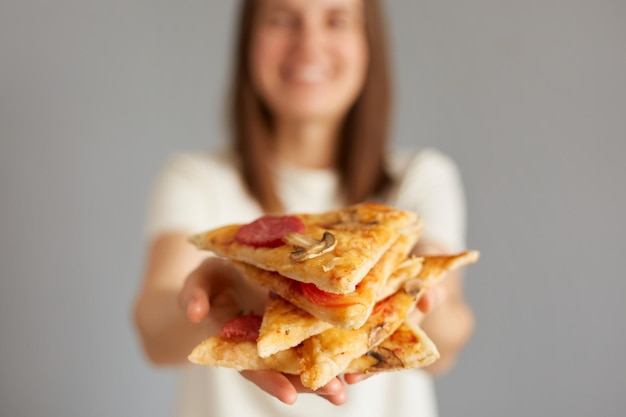 ピザのスライスを保持している満足している女性の写真は、ピッツェリアで喜んでいると感じており、灰色の背景に分離されたカジュアルな服装を着てジャンク フードを食べることを申し出てカメラを直接見て喜んでいます