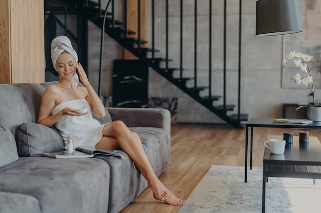 Photo of satisfied young European woman sits on comfortable sofa in living room applies face cream for reducing wrinkles wrapped in bath towel reads magazine undergoes beauty treatments at home