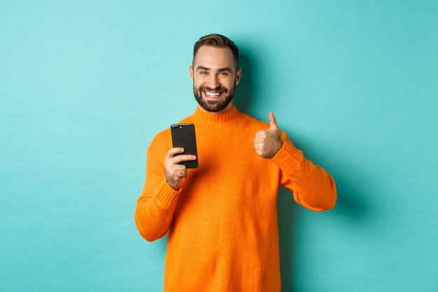 Photo of satisfied young man in orange sweater, showing thumb-up after reading on mobile phone, standing pleased against turquoise background.