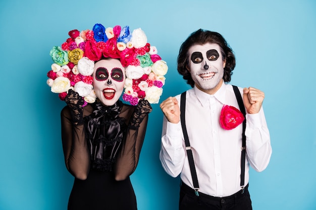 Photo of scary two people man lady surprised raise fists win undead role play character competition wear black dress death costume roses headband suspenders isolated blue color background