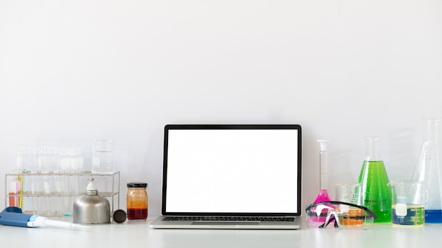 白い空白の画面のコンピューターラップトップで白い作業机に置く科学実験装置の写真。フラット横たわっているコンピューターのラップトップ、化学ガラス製品、安全メガネ。