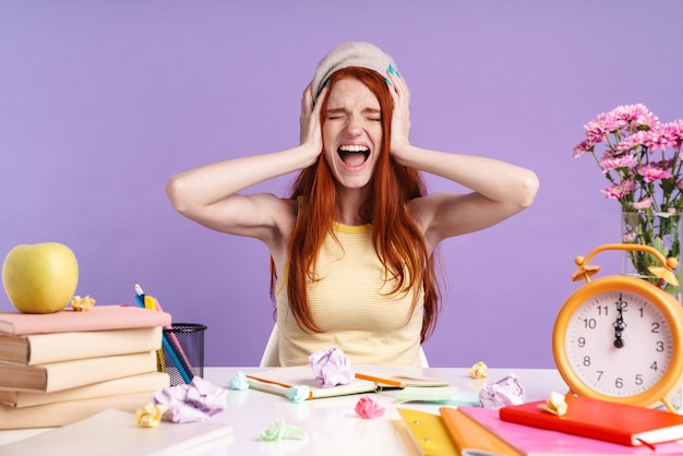 Photo photo of screaming student girl grabbing her head while sitting at desk with exercise books isolated over purple wall