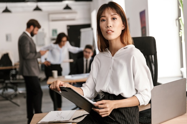 Photo of serious asian businesswoman looking at front and holding clipboard in open-plan office