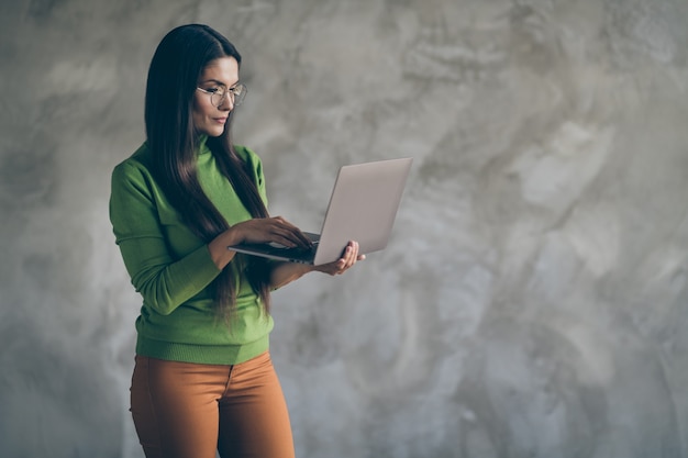 필요한 정보를 찾기 위해 입력하는 주황색 바지에 노트북을 통해 검색 심각한 자신감 스마트 영리한 여자의 사진 격리 된 회색 벽 색 콘크리트 배경