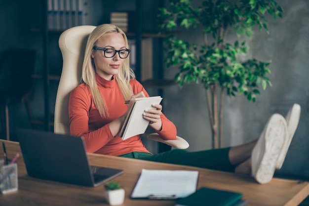 La foto della donna sicura seria che esamina il taccuino che scrive i suoi pensieri doveva essere utilizzata in avvio con i piedi sul desktop