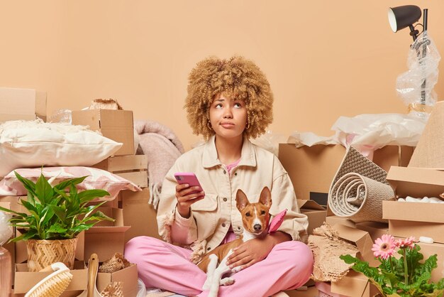 Photo of serious curly haired woman focused above being deep in
thoughts holds mobile phone with dog thinks about message content
relocates to new house packs things in boxes real estate
concept