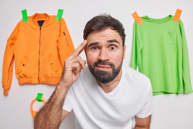 Photo of serious handsome bearded European man concentrated at camera keeps finger on temple tries to remember something poses against items of clothing hanging on white wall has bad memory.