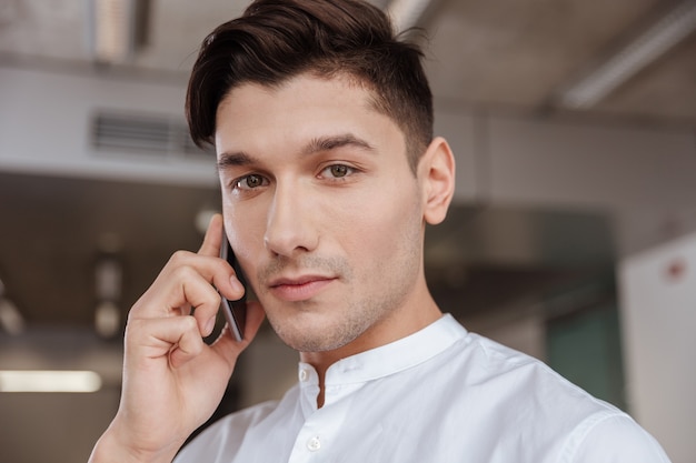 Photo of serious man dressed in white shirt talking by the phone indoors. Coworking. Looking at camera.