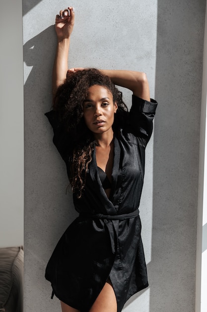 Photo of sexual african american woman wearing housecoat, posing over concrete wall indoor