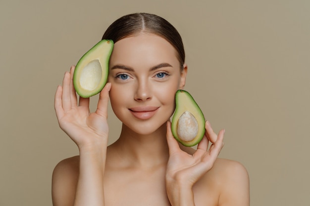 Photo of shirtless female model looks with gentle expression, holds halves of avocado near face has healthy well cared skin after cosmetic treatments