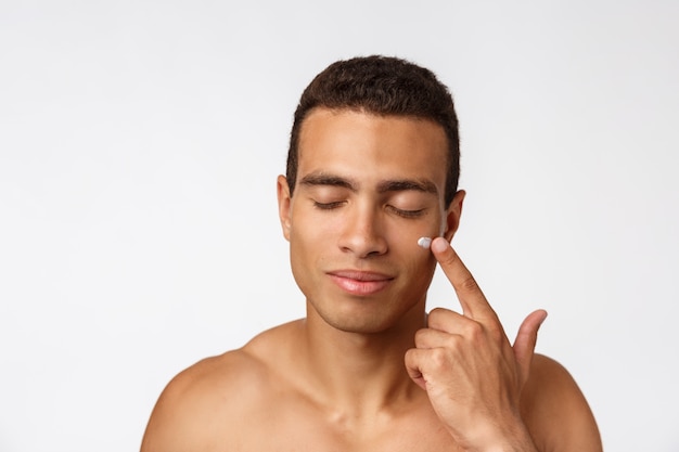 Photo of shirtless man smiling and applying face cream isolated over white