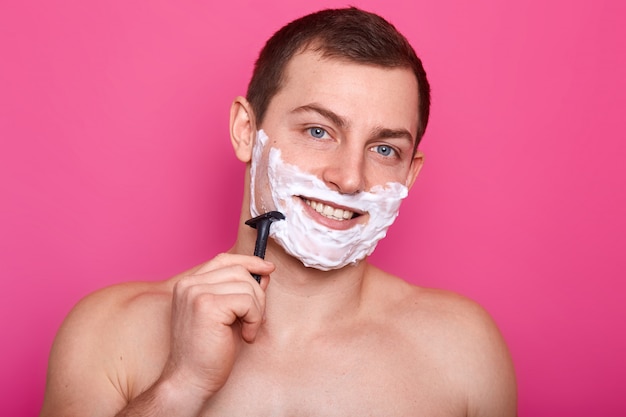 Photo of shirtless young man shaving his face and looking at camera, posing naked with smile