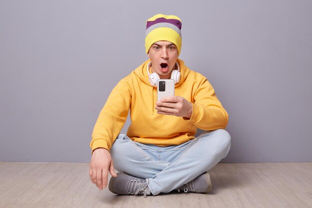 Photo of shocked amazed young adult man wearing beanie hat yellow hoodie and jeans posing against gray wall sitting on floor with crossed legs using mobile phone looking at screen with big eyes