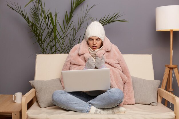 Photo of shocked surprised astonished chilly woman wearing cap and winter coat sitting on the sofa at home working on laptop looking at display with big eyes