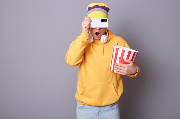 Photo of shocked surprised unknown man wearing yellow hoodie and beanie hat posing in cinema against gray wall holding paper bucket with popcorn covering his eyes with smart phone hiding