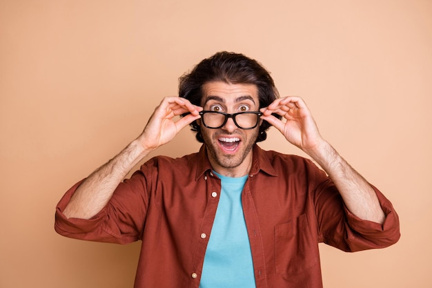 Photo of shocked young guy hands hold eyeglasses look camera wear specs brown t-shirt isolated beige color background