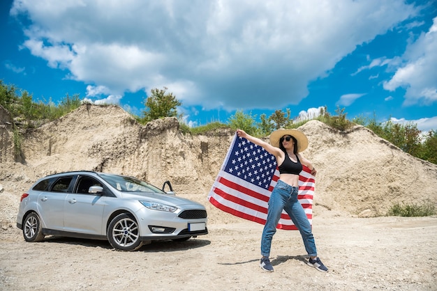 夏の砂の採石場で車の近くにアメリカ国旗を持つ魅力的な女性の写真撮影