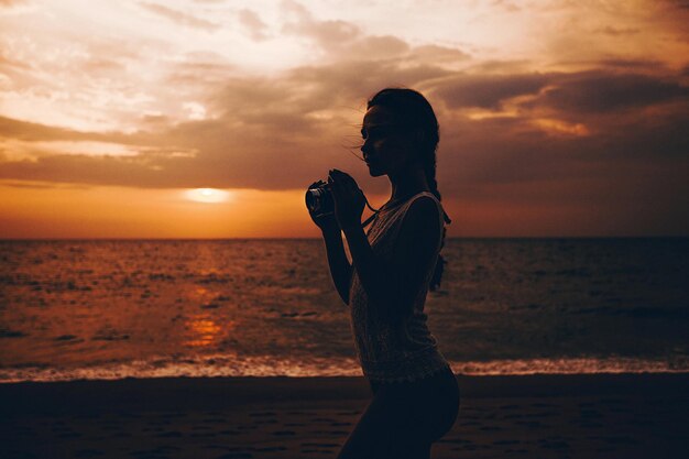 Photo shoot at sunset. Young beautiful girl in lace top and straw hat stands by the ocean with a camera in her hands and looks at the landscape. Tropical landscape with views of the sea and the coast.