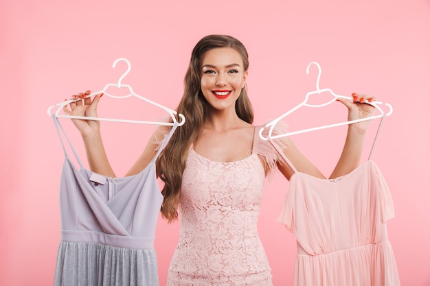 Photo of shopaholic woman 20s holding and choosing two dresses on hangers while shopping, isolated over pink wall