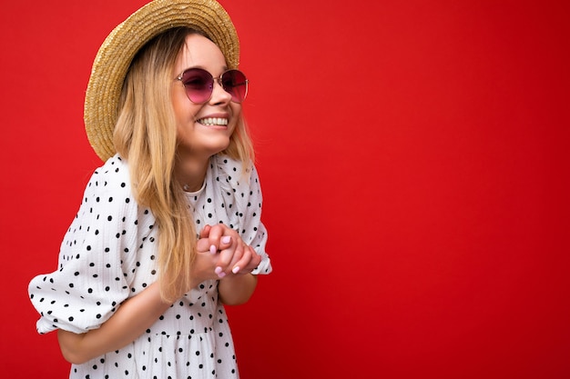 夏のカジュアルな服とスタイリッシュなサングラスを身に着けている魅力的な幸せな笑顔の若いブロンドの女性の写真撮影は、横を向いているカラフルな背景の壁の上に分離されました。コピースペース