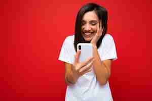 Photo photo shot of attractive positive surprised good looking young brunette woman wearing casual stylishwhite t-shirt poising isolated on red background with empty space holding in hand and using mobile p