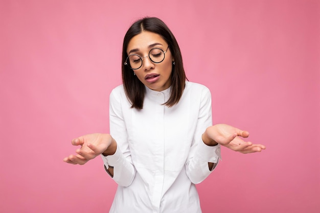 Photo shot of beautiful asking sad young brunette woman wearing casual clothes and stylish optical glasses isolated over colorful background and having questions