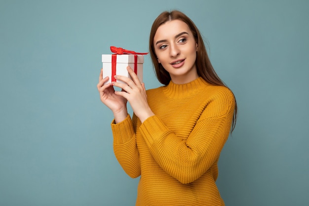 photo shot of beautiful happy brunette young woman isolated over blue background wall wearing mustard sweater holding white gift box with red ribbon and looking to the side.
