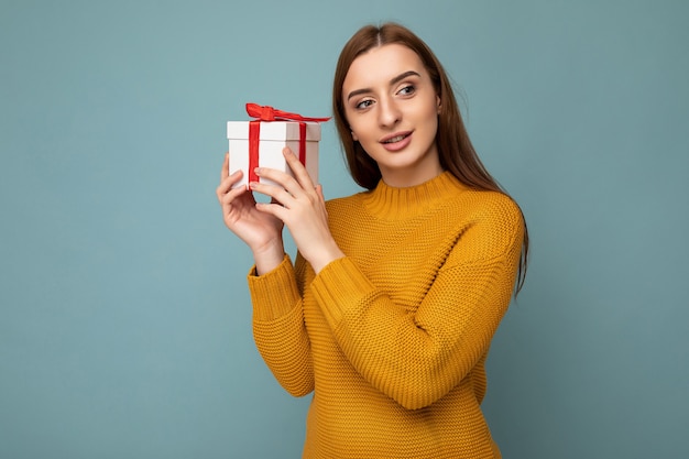 Photo shot of beautiful happy brunette young woman isolated over blue background wall wearing mustard sweater holding white gift box with red ribbon and looking to the side.