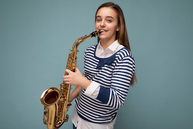 Photo shot of beautiful happy smiling brunette little girl wearing striped longsleeve standing isolated over blue background wall playing saxophone looking at camera.