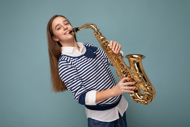 Photo shot of beautiful happy smiling brunette little girl wearing striped longsleeve standing isolated over blue background wall playing tube musical instrument looking at camera