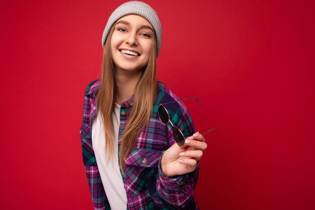 Photo shot of beautiful positive smiling young brunette woman wearing summer casual clothes and holding stylish sunglasses isolated over colorful background wall looking at camera.