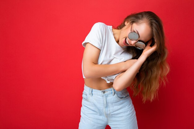 Photo shot of beautiful young positive dark blonde woman wearing casual clothes and stylish sunglasses isolated over colorful background and having fun