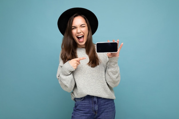 Scatto fotografico della giovane e bella donna che indossa cappello nero e maglione grigio tenendo il telefono che mostra lo smartphone isolato su sfondo.mock up, ritaglio, spazio di copia