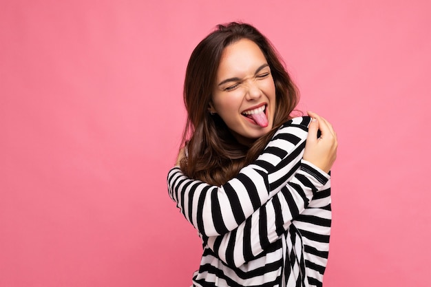 Photo shot of cute attractive pretty young happy joyful brunette woman wearing casual striped longsleeve isolated over colorful background with copy space and showing tongue.