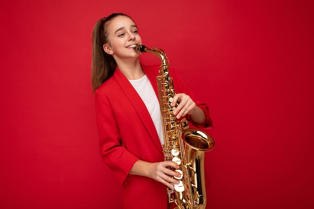 Foto scattata di una bambina bruna sorridente abbastanza felice che indossa una giacca rossa elegante in piedi isolata su una parete di fondo rossa che suona il sassofono guardando di lato.