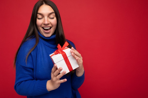 Photo shot of pretty positive surprised young brunet woman isolated over colourful background wall wearing trendy outfit look holding gift box and looking at present box with red ribbon. Free space