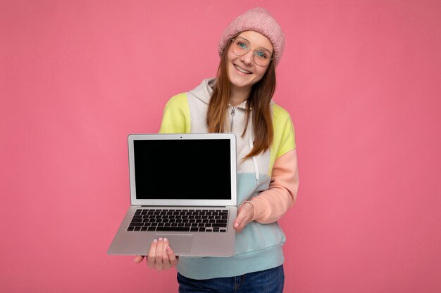 Photo shot of smiling Pretty young lady wearing hat sweater and glasses holding computer laptop wearing white headphones looking at camera isolated over pink background. Mock up