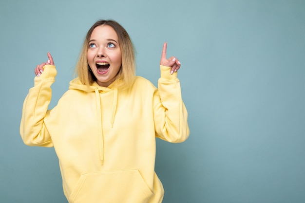 Photo shot of young beautiful cute happy blonde woman wearing stylish yellow hoodie isolated over