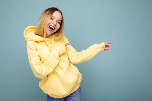 Photo shot of young beautiful cute happy blonde woman wearing stylish yellow hoodie isolated over