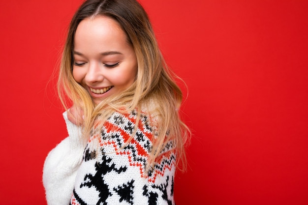 Photo shot of young beautiful cute happy blonde woman wearing winter sweater isolated over red