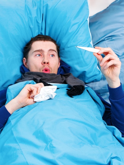 Photo of sick brunet man in scarf with thermometer in hand lying in bed