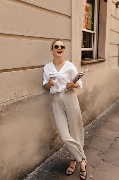 Photo slim caucasian blond woman looks to side through sunglasses standing on street with coffee